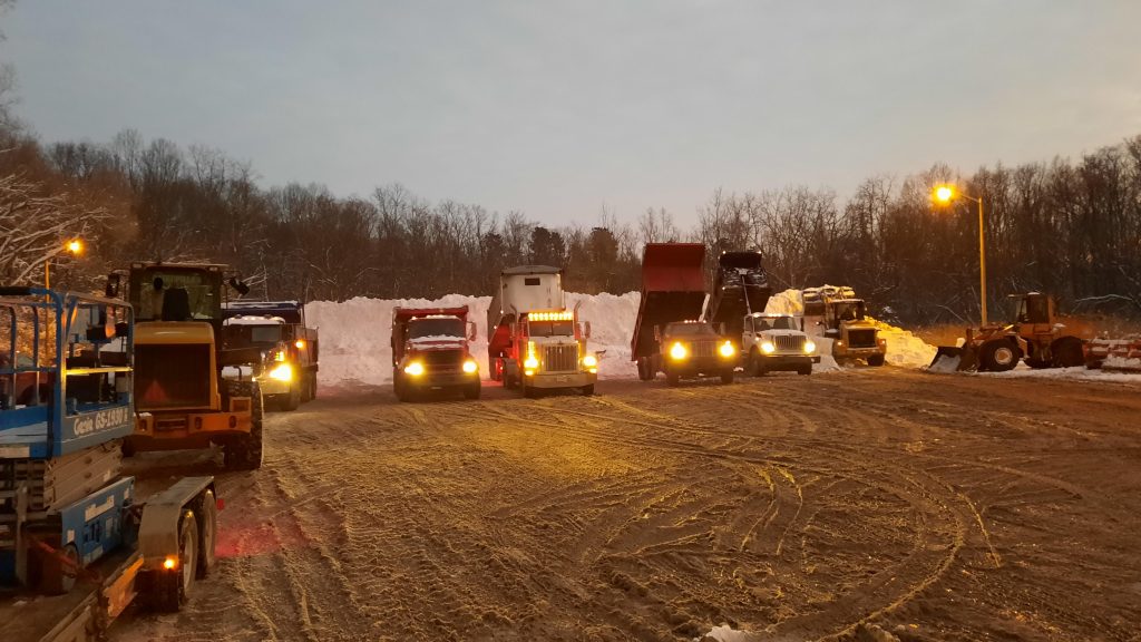 5 dump trucks backed up to a snow pile in semi-darkness