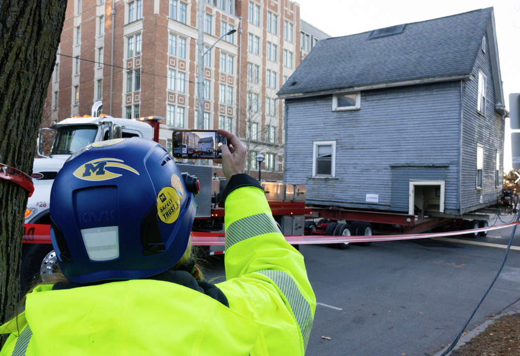 A worker wearing a high-visibility jacket and blue safety helmet photographs a gray house being transported on a flatbed truck along a city street, with a multi-story brick building in the background.