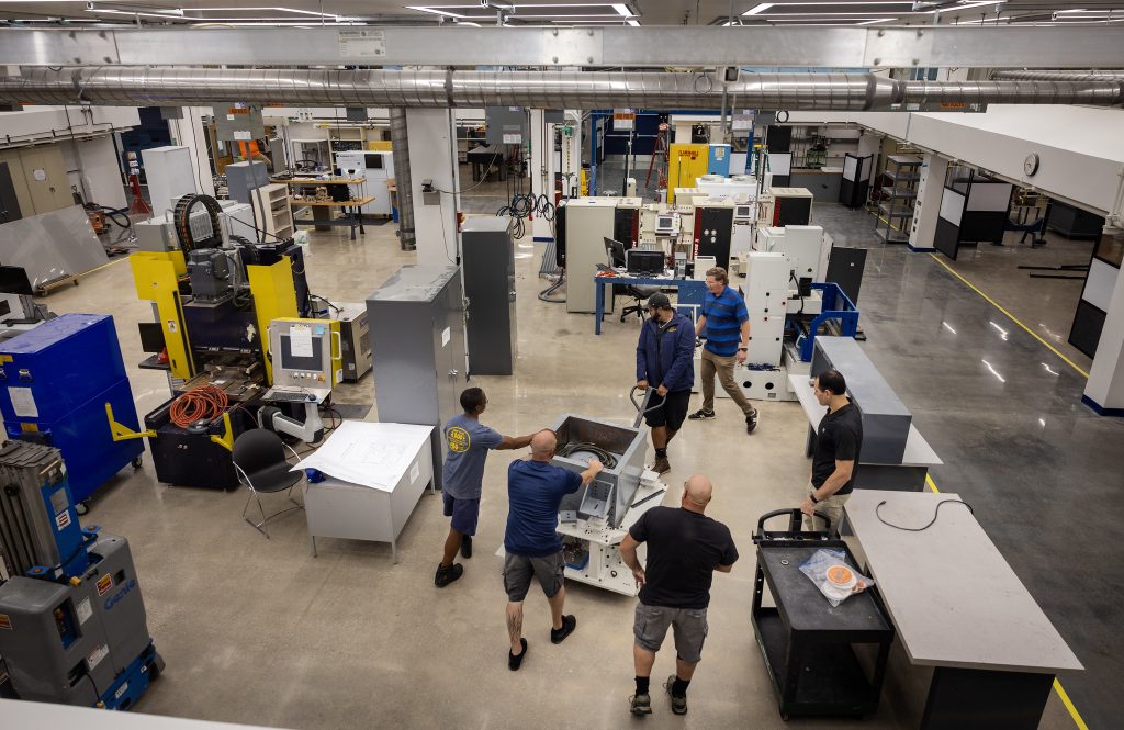 Moving and trucking crew members position heavy equipment inside a large, upgraded manufacturing lab space (Dow 1100) at the Dow Building on the University of Michigan’s North Campus. The bright, industrial room features high ceilings, exposed ductwork, machinery, worktables, and tools as workers maneuver a large metal unit across the polished concrete floor.