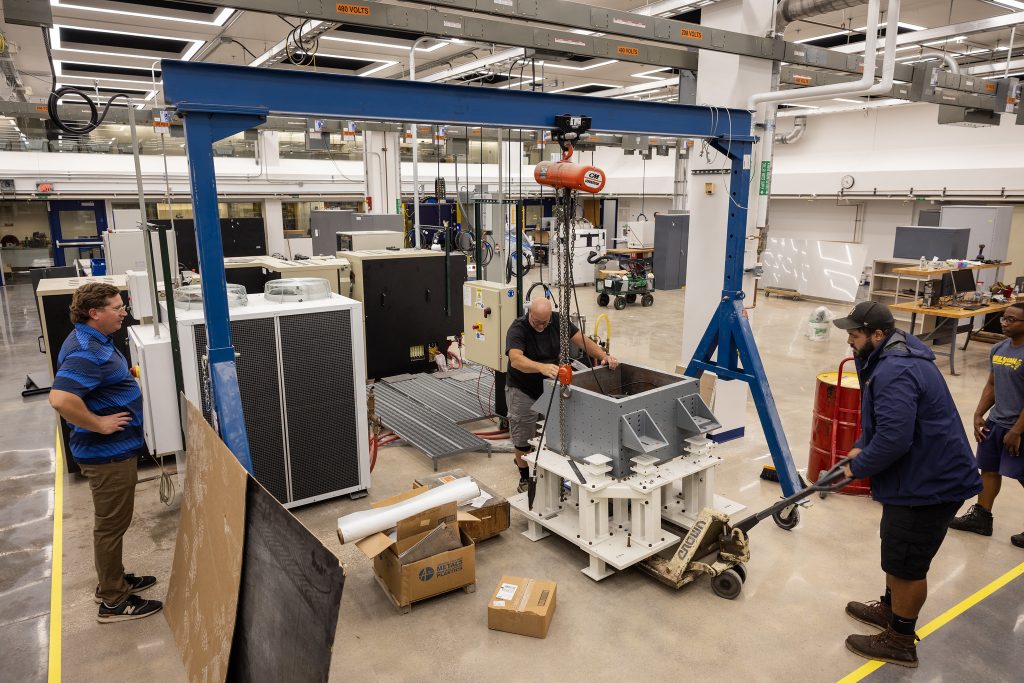 Workers use an overhead hoist to move a heavy machine component, with several people guiding and observing the lift. From left are: Matt Jastrzembowski, Dow Building Manager, then Joe McGregor, Dante Barnett and Donovan Miles from Moving and Trucking.