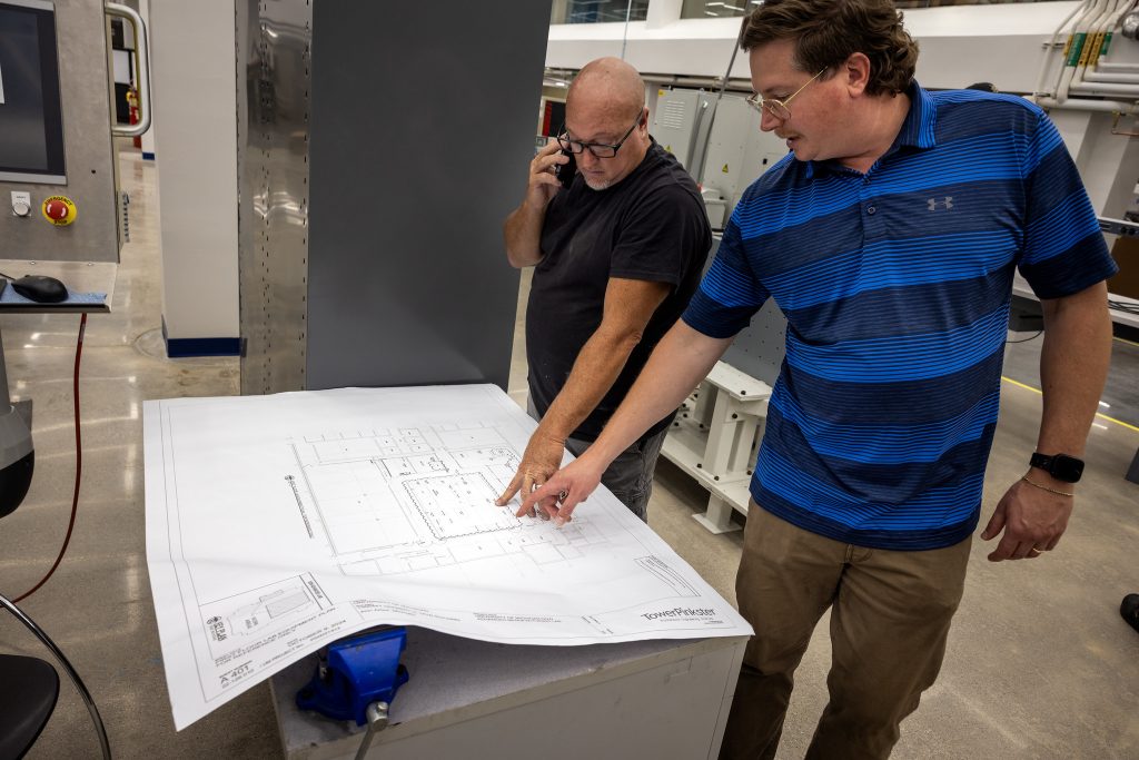 Matt Jastrzembowski, Dow Building Manager (right) and Joe McGregor review and point at a large technical blueprint spread out on a table in an industrial workspace.