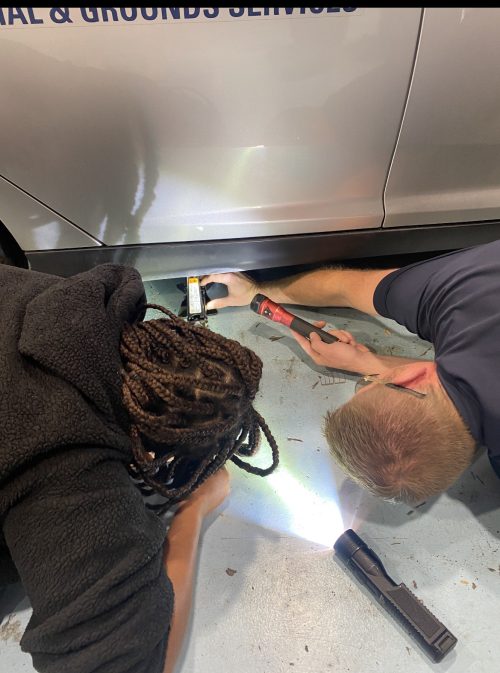 A student and a mechanic fly on the floor looking at a car jack with a flashlight.